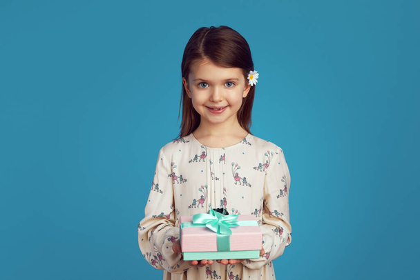 Маленькая девочка демонстрирует завернутую подарочную коробку с лентой на синей стене - Фото, изображение