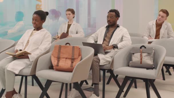 Повільний знімок багатоетнічної групи молодих веселих студентів-медиків у білих халатах, що сидять на стільцях у сучасній аудиторії, аплодуючи професору після цікавої лекції
 - Кадри, відео