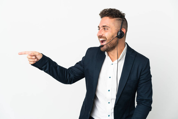 Homem telemarketer trabalhando com um fone de ouvido isolado no fundo branco apontando o dedo para o lado e apresentando um produto - Foto, Imagem