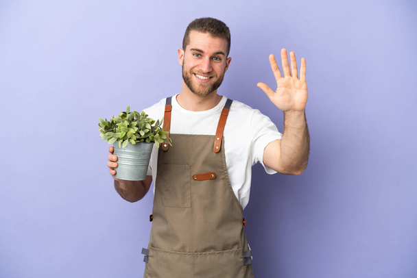 Jardinier homme caucasien tenant une plante isolée sur fond jaune saluant à la main avec expression heureuse - Photo, image