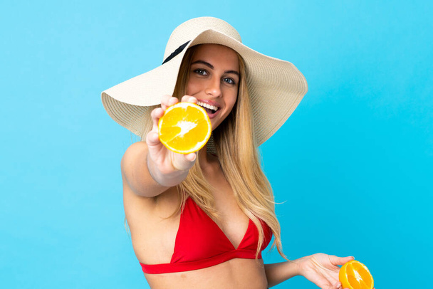 Young Uruguayan blonde woman over isolated blue background in swimsuit and holding an orange - Foto, Imagen