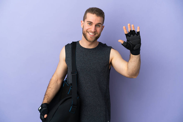 Mladý sportovní muž se sportovní taškou izolované na bílém pozadí salutování s rukou s veselým výrazem - Fotografie, Obrázek