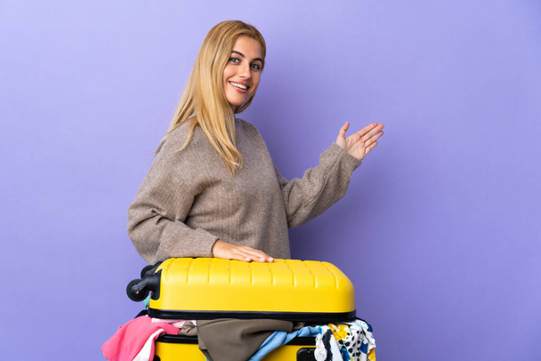 Mladá uruguayská blondýnka s kufříkem plným šatů přes izolovanou purpurovou stěnu natahující ruce na stranu, aby ji pozvala - Fotografie, Obrázek