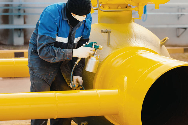 A painter in working clothes paints a metal shut-off valve for gasification from a compressor gun on a summer day.  - Photo, Image