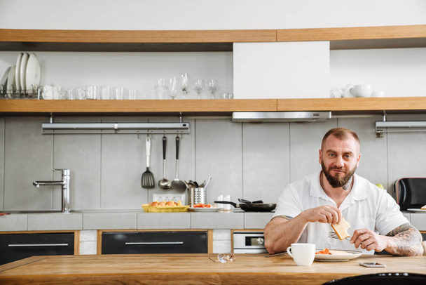 White man with beard having lunch while sitting at table in kitchen - Foto, Imagem
