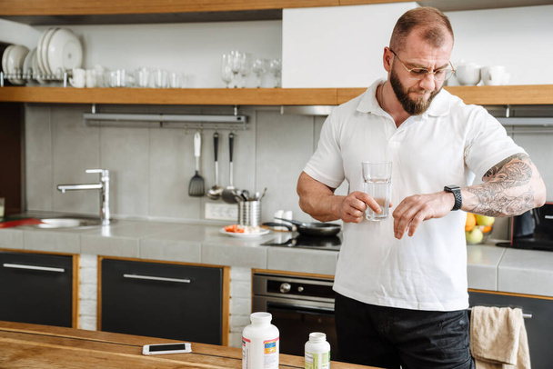 White man in eyeglasses looking at wristwatch while taking her medicine in kitchen - Фото, зображення