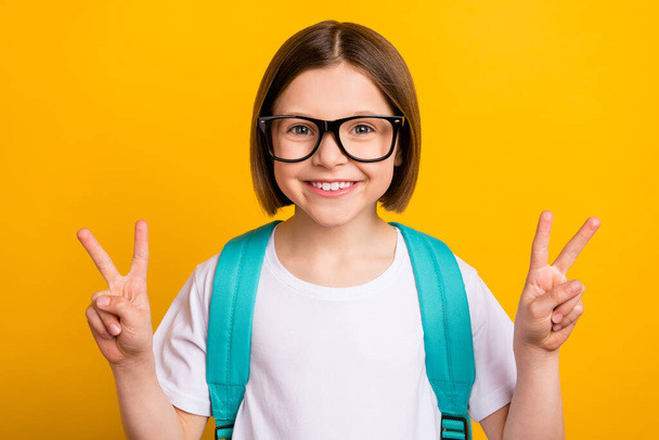 Photo portrait small schoolgirl smiling wearing glasses showing v-sign gesture isolated vivid yellow color background - Fotó, kép