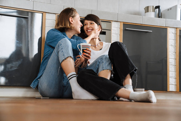 A smiling lesbian couple looking to each other while sitting on the floor with a cup in the light kitchen - Foto, imagen