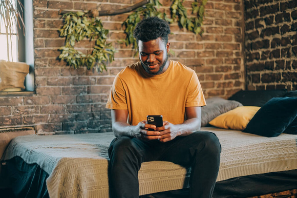 Black man texting on his mobile phene sitting on the bed indoors at home - Φωτογραφία, εικόνα
