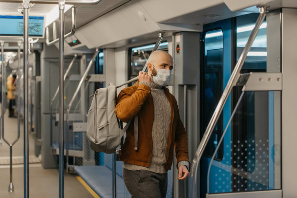 Een man met een baard in een gezichtsmasker om de verspreiding van het coronavirus te voorkomen zet een grijze rugzak op terwijl hij met de metro rijdt. Een kale man met een chirurgisch masker houdt sociale afstand op een trein.. - Foto, afbeelding