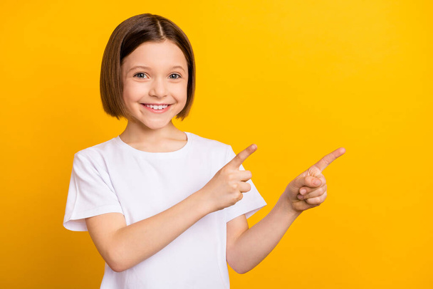 Photo of charming sweet school girl wear white t-shirt smiling pointing two fingers empty space isolated yellow color background - Фото, зображення