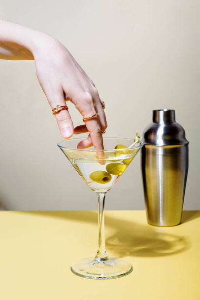 Woman dips finger with rings in glass of martini cocktail with olives - Φωτογραφία, εικόνα