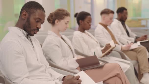 Πλάγια εικόνα ομάδας νεαρών πολυεθνικών φοιτητών ιατρικής με λευκά παλτά που κάθονται στη σειρά στη σύγχρονη τάξη ακούγοντας διαλέξεις και κρατώντας σημειώσεις - Πλάνα, βίντεο
