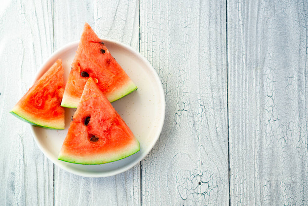 Triangular slice of cool watermelon on a round white plate on the blue surface of the kitchen table. Freshness in February heat with vitamins - Fotografie, Obrázek