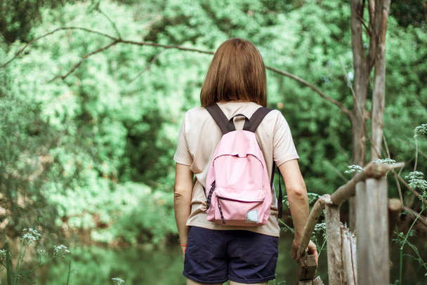 A young woman with short hair and a pink backpack stands with her back to the camera on a small wooden bridge near a river in the woods. Selective Focus. - Photo, Image