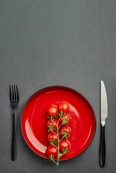 Ντομάτες σε ένα πιάτο ως δίαιτα ή υγιεινά τρόφιμα έννοια. Ντομάτες σε κόκκινο πιάτο με πιρούνι και μαχαίρι σε μαύρο πέτρινο φόντο. Θέση για κείμενο ή σχεδιασμό - Φωτογραφία, εικόνα