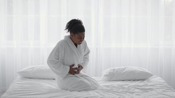 Female period pain. Young sick african american woman wearing bathrobe suffering from abdominal pain, sitting on bed - Footage, Video