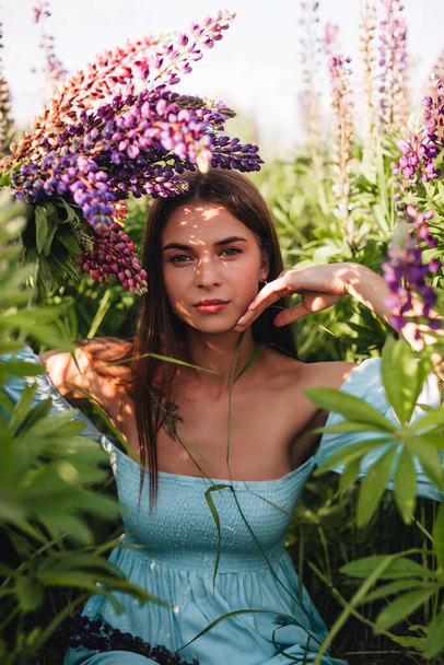 A beautiful girl with long hair in a blue dress holds a bouquet of lupines in a field of flowers. - Photo, Image