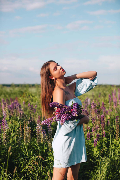 Een mooi meisje met lang haar in een blauwe jurk houdt een boeket lupine in een veld van bloemen. - Foto, afbeelding
