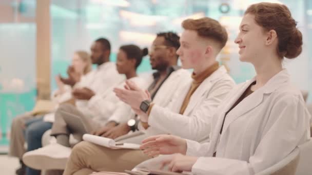 Vista lateral de plano medio de grupo de jóvenes estudiantes de medicina multiétnica en batas blancas aplaudiendo al profesor sentado en fila en la moderna sala de conferencias - Imágenes, Vídeo