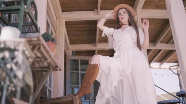 Rapariga do campo no alpendre, vestido branco e chapéu de cowboy. Acção. Visão inferior de um modelo feminino posando com a perna na bota de cowboy marrom em uma mesa de madeira. - Filmagem, Vídeo