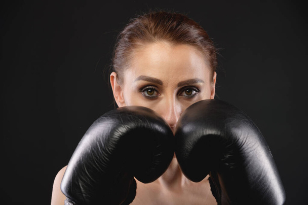 De jonge blanke sportvrouw in sportkleding en grote zwarte bokshandschoenen kijkt in de camera en bedekt haar gezicht met handschoenen. Kopieerruimte - Foto, afbeelding