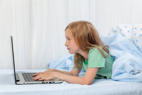 Девушка лежит дома на кровати под одеялом с ноутбуком. Дистанционное обучение. Компьютерные игры. Уроки. Домашнее образование. - Фото, изображение