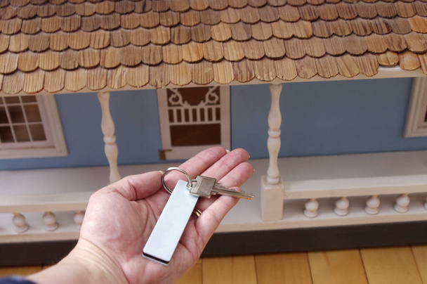 mano sosteniendo las llaves de plata con etiqueta en frente de la casa de muñecas de madera azul grande  - Foto, imagen