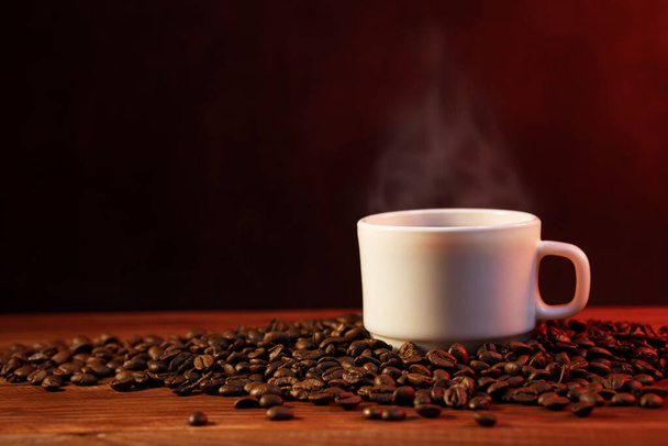 Warm cup of coffee and coffee beans on dark background. Coffee cup and coffee beans on wooden table. - Zdjęcie, obraz