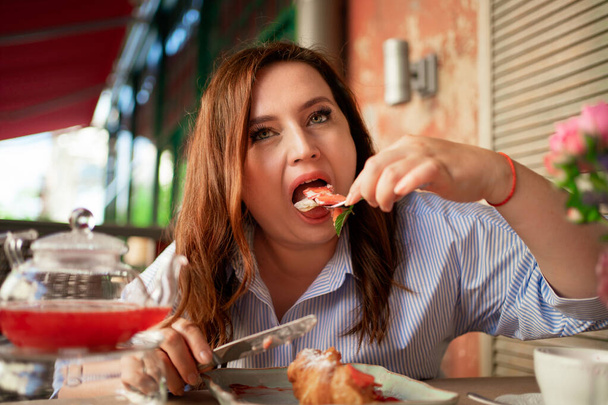 gluttony. A funny woman eats a croissant with strawberries with a knife and fork - Foto, Imagem