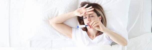 Η άρρωστη γυναίκα ξαπλώνει στο κρεβάτι με το θερμόμετρο από κοντά. - Φωτογραφία, εικόνα