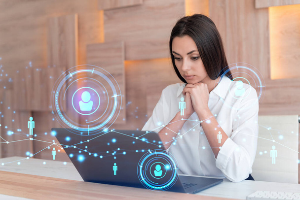 Attractive businesswoman in white shirt at workplace working with laptop to hire new employees for international business consulting. HR, social media hologram icons over office background - Foto, Imagem