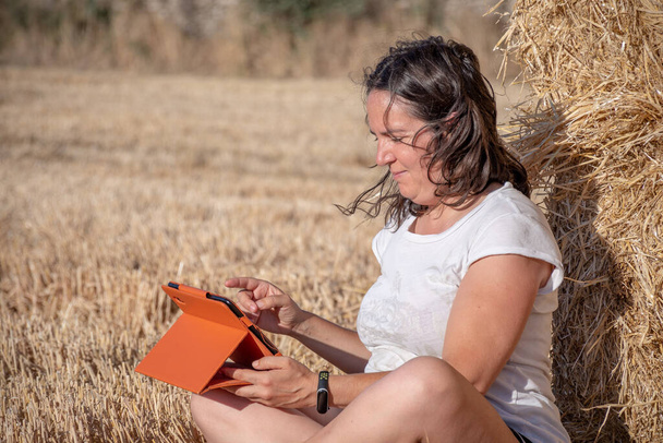 Orta yaşlı esmer Latin kadın bir tarlada oturup saman balyalarına yaslanarak turuncu kapaklı tablet kullanıyor. Teknolojik ve kırsal bölgeler. - Fotoğraf, Görsel