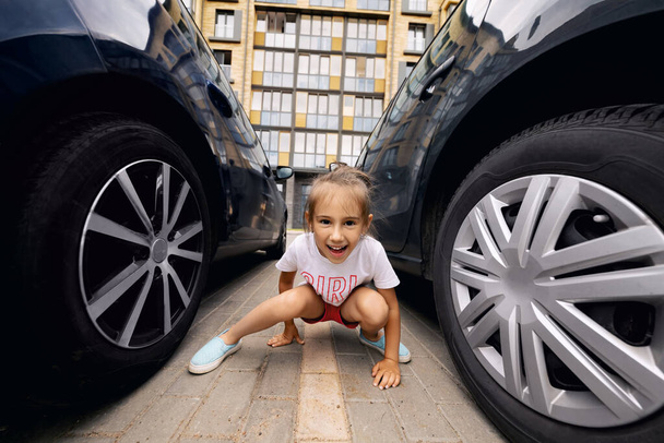 Ein Mädchen sitzt an einem warmen Sommertag auf den Fahrbahnmarkierungen auf dem Parkplatz zwischen zwei Autos. Ein mehrstöckiges Haus im Hintergrund hinter einem Kind - Foto, Bild