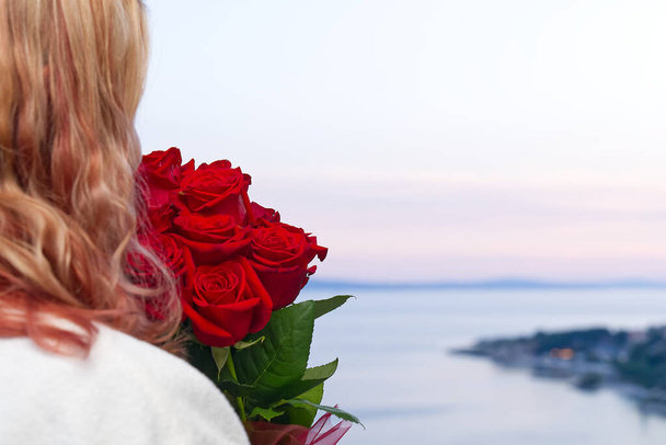 Glückliche Frau in weißer Robe mit einem Strauß roter Rosen blickt vom Hotelbalkon auf das Meer - Foto, Bild