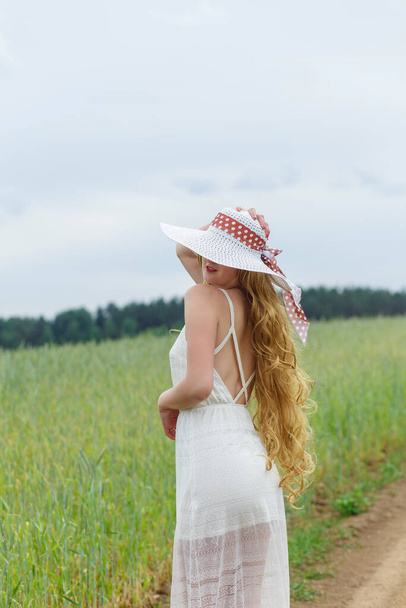 Girl in a white dress and hat on a field background. The girl in a hat stands, relaxing outdoors, having fun, harmony concept. - Zdjęcie, obraz