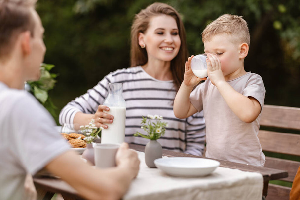 Colazione in famiglia nella natura in giardino. Allegro mamma e piccolo figlio con papà bere latte e mangiare biscotti freschi in una mattina d'estate a tavola - Foto, immagini