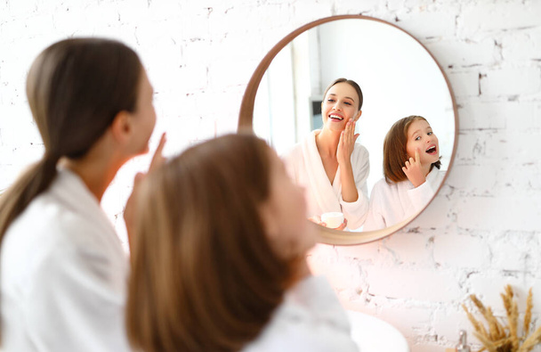 Happy young woman with preteen daughter standing near mirror and applying skin care product on face while enjoying beauty procedure together at home - Photo, Image