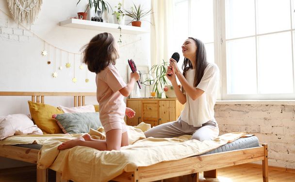 Zwei glückliche Mädchen, die sich als berühmte Sängerinnen ausgeben, überglückliche jüngere und ältere Schwester mit Haarbürste als Mikrofon singen, während sie auf dem Bett sitzen. Jung mutter und süß klein tochter having spaß bei zuhause - Foto, Bild