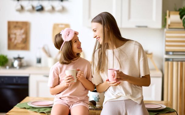 Szczęśliwy czas z mamą. Młoda piękna matka i urocza córeczka pijąc herbatę i uśmiechając się stojąc razem w nowoczesnej, przytulnej kuchni, starsza i młodsza siostra ciesząca się rodzinnym weekendem w domu - Zdjęcie, obraz