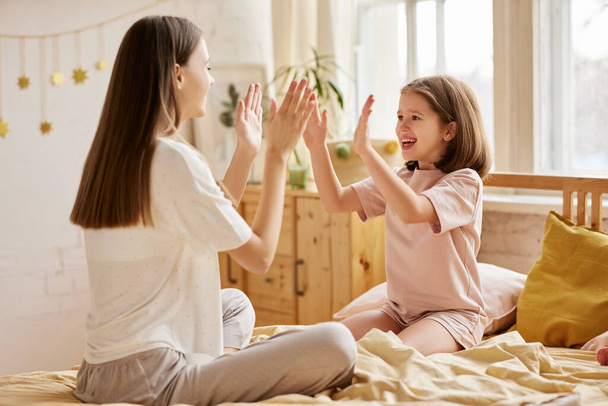 Enjoying time with family. Two caucasian girls, younger and older happy sisters in pajamas playing together while sitting on bed in the morning, young mom and cute little daughter having fun at home - Photo, Image