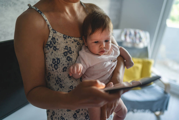 Dorosła kobieta matka gospodarstwa trzymiesięczne dziecko podczas korzystania z telefonu komórkowego do przeglądania Internetu lub wiadomości w sieci społecznej w pokoju w domu życie rodzinne macierzyństwo i rodzicielstwo zajęty koncepcji - Zdjęcie, obraz