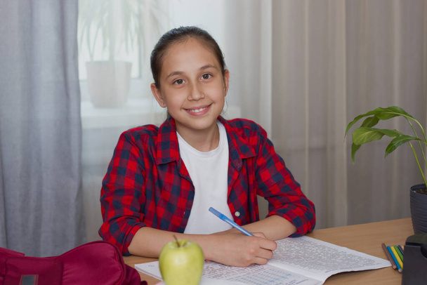 Девушка с темными длинными волосами в белой футболке в красной рубашке в клетке сидит за столом, пишет, делает домашнее задание, занимается. Смотрит в камеру. Снова в школу - Фото, изображение