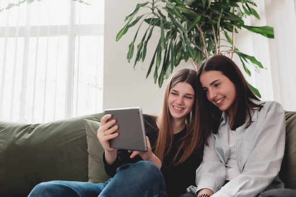 Twee tienermeisjes zitten op de groene bank in de woonkamer en kijken naar een grappige video op tablet, glimlachend, lachend. Mooie mensen, frendship en saamhorigheid concept. Bloempot op rug - Foto, afbeelding