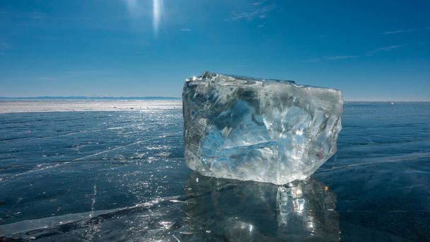 Un cristal de hielo transparente se encuentra en la superficie lisa y azul de un lago congelado. Deslumbramiento del sol en los bordes, reflejo. Cielo azul claro, día de invierno soleado. Primer plano. Baikal. - Foto, Imagen