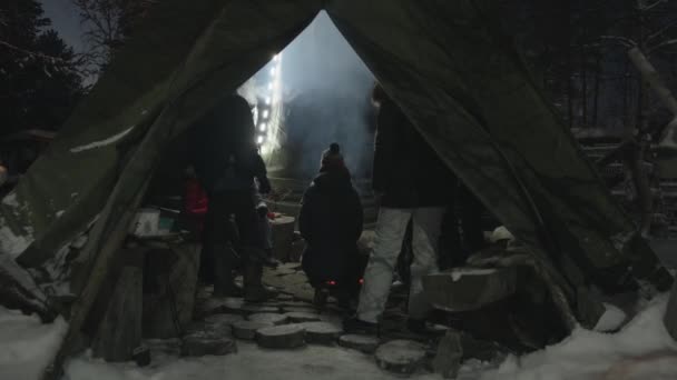 Un grupo de turistas se sienta en un wigwam en un patio cubierto de nieve alrededor del hogar con un fuego y se comunica - Metraje, vídeo