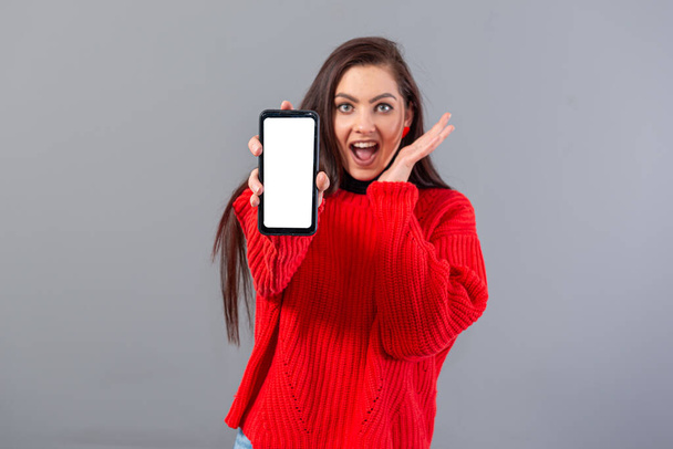 surprised young teenager woman dressed in red sweater shows her smartphone, isolated on gray - Fotografie, Obrázek
