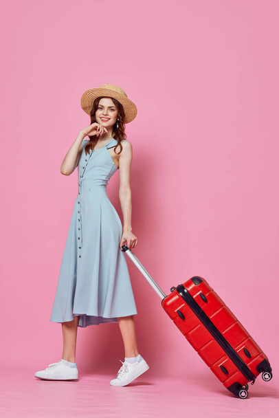 γυναίκα σε μπλε φόρεμα με κόκκινη βαλίτσα ταξιδιωτικό προορισμό ροζ φόντο - Φωτογραφία, εικόνα