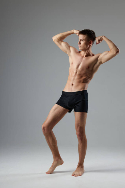 athlete in black shorts holds hands behind his head naked torso gray room full-length - Photo, Image