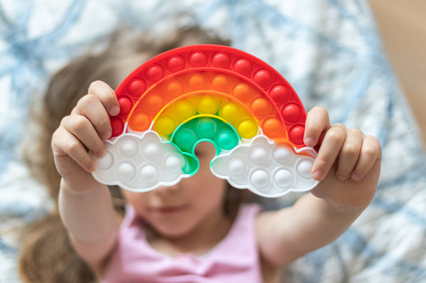 Klein blond meisje houdt een speelgoed pop it in de vorm van een regenboog in haar handen - Foto, afbeelding